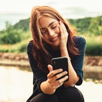 Liebeserklärung Whatsapp: Frau mit braunen Haaren sitzt in der Natur an ihrem Handy und lacht