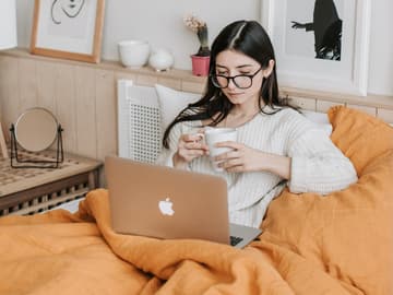 Mädchen im Bett mit einer Tasse in der Hand und Laptop auf dem Schoß