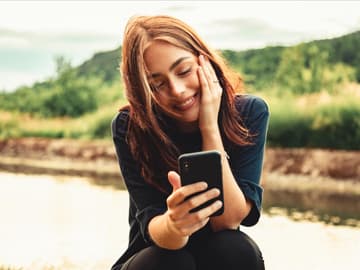 Liebeserklärung Whatsapp: Frau mit braunen Haaren sitzt in der Natur an ihrem Handy und lacht