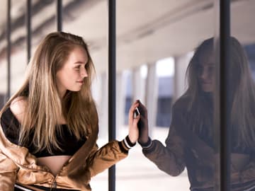 Wer bin ich? Junges Mädchen mit blonden Haaren schaut auf ihre Spiegelung in einem Fenster
